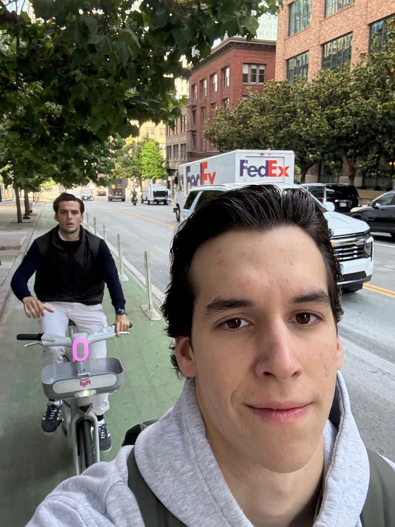 A selfie of Marcos on a city street, with a cyclist behind him
